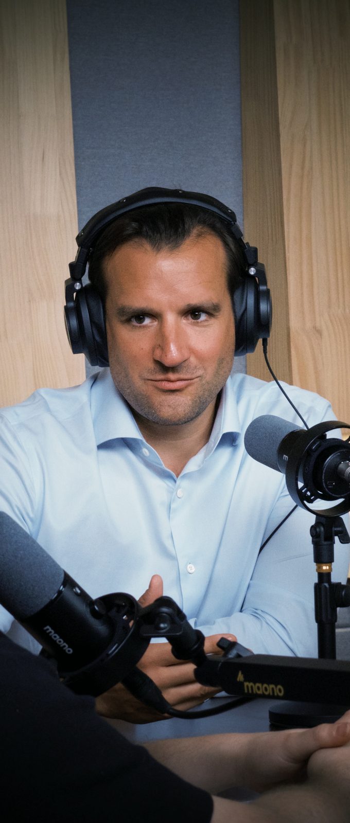 Rafael Ratzel at the mic with studio headphones, light blue shirt, looking straight into the lens, the moment he describes lifelong friends accusing him of being brainwashed at European dinners
